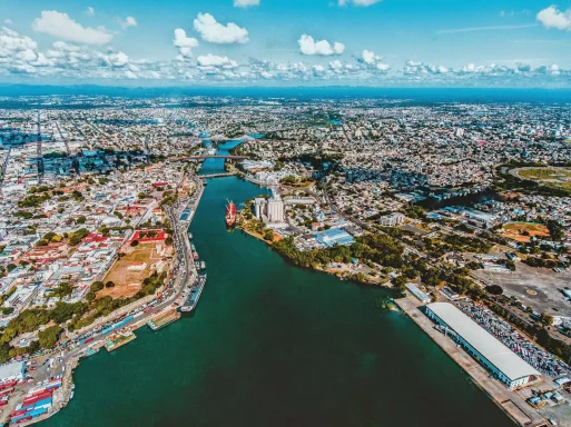 SANTO DOMINGO - Einw. 3.172.000 Agglomeration Luftaufnahme einer Stadt mit Fluss, Gebäuden und grünen Flächen unter einem blauen Himmel.